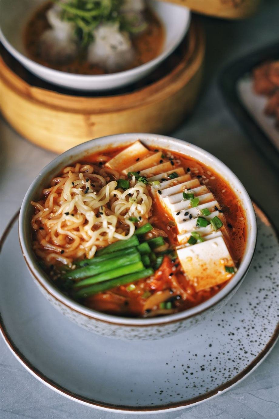 A bowl of kimchi ramen with tofu, scallions and sesame, beside a bamboo steamer of dumplings